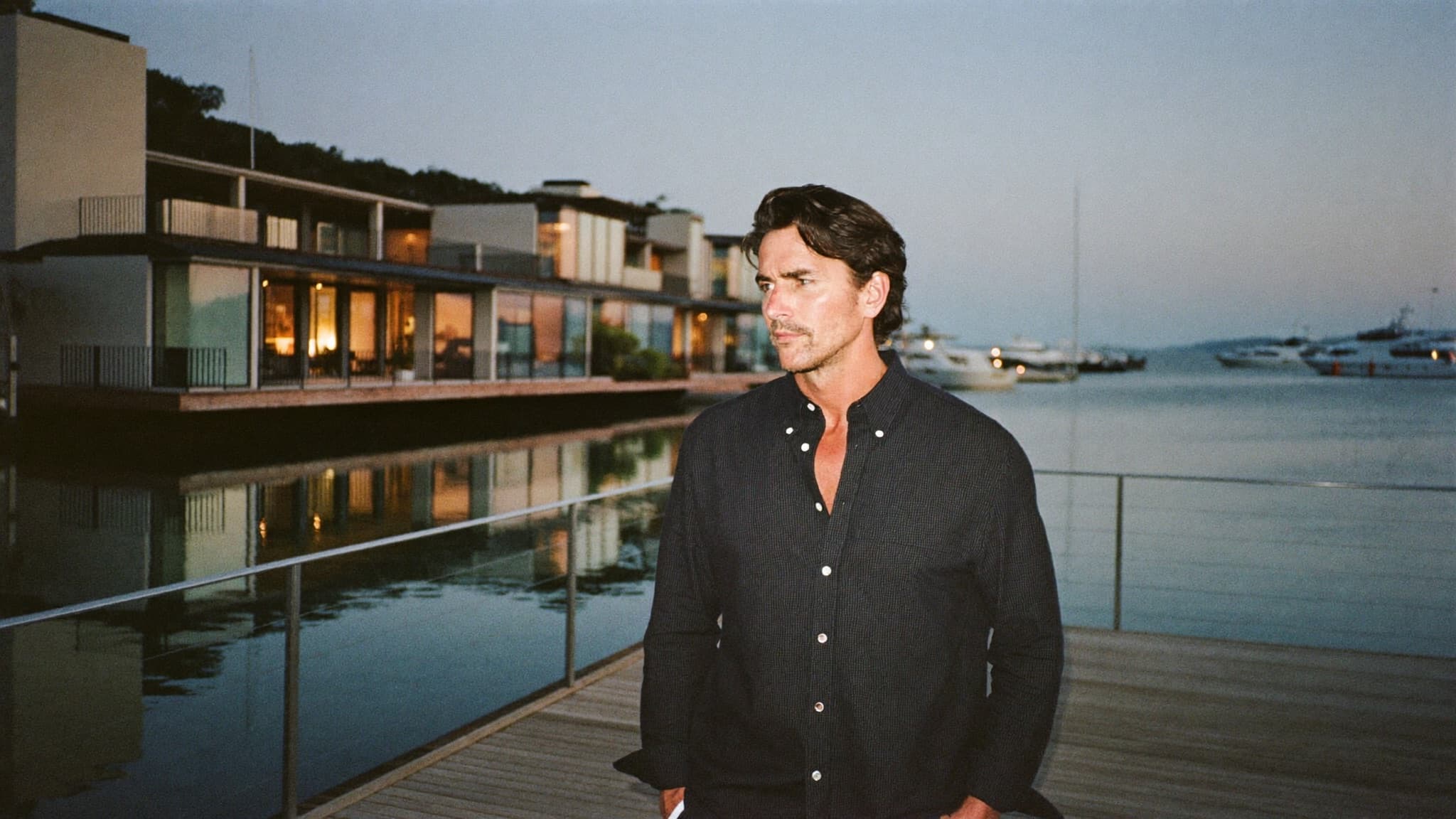 Justin Ratowsky standing on a Huntington Harbour dock at twilight, waterfront homes glowing behind calm water, Ratowsky Group at Compass luxury waterfront specialists.