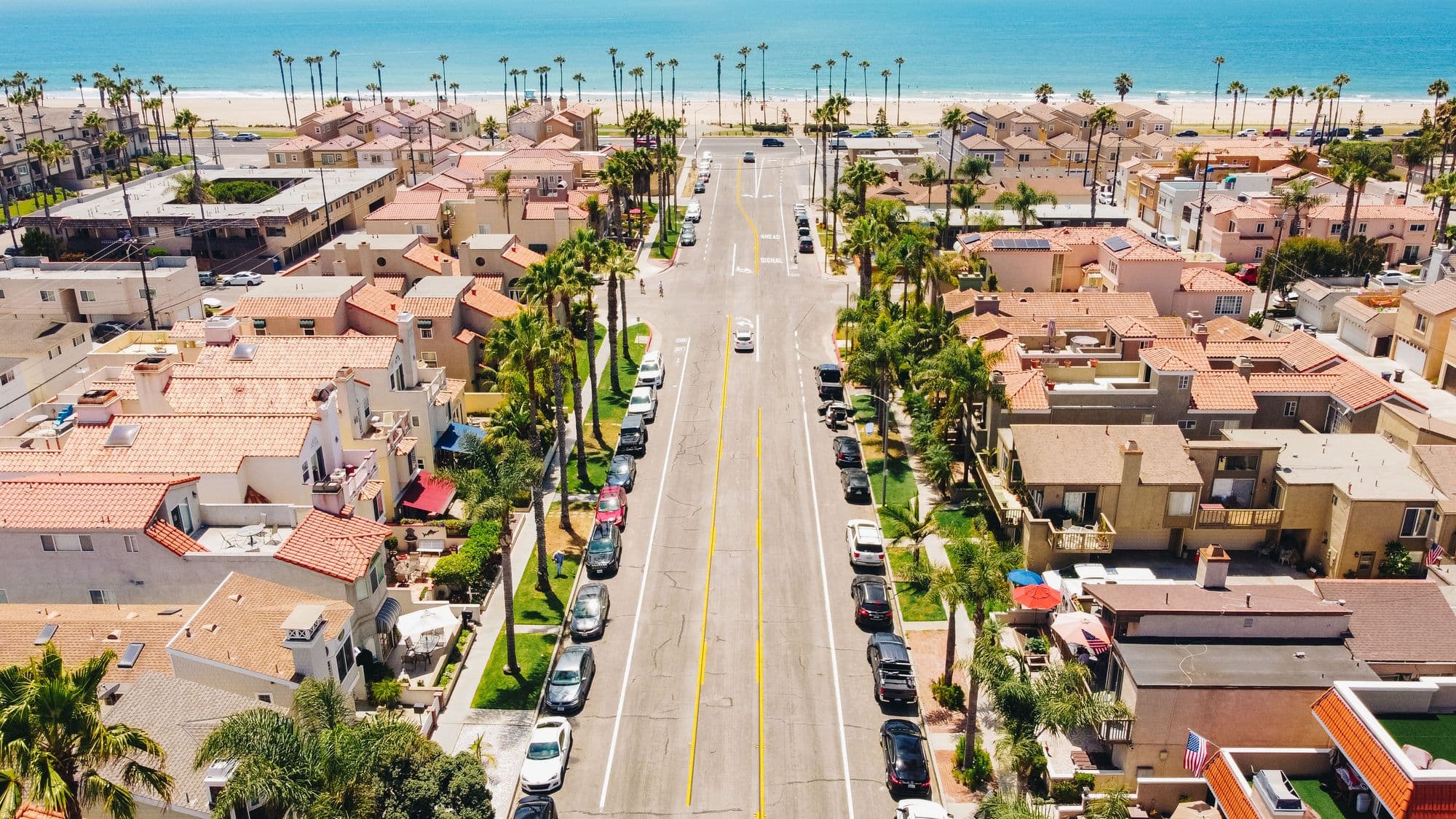 Aerial of a palm-lined downtown Huntington Beach street running straight to the sand near the pier.