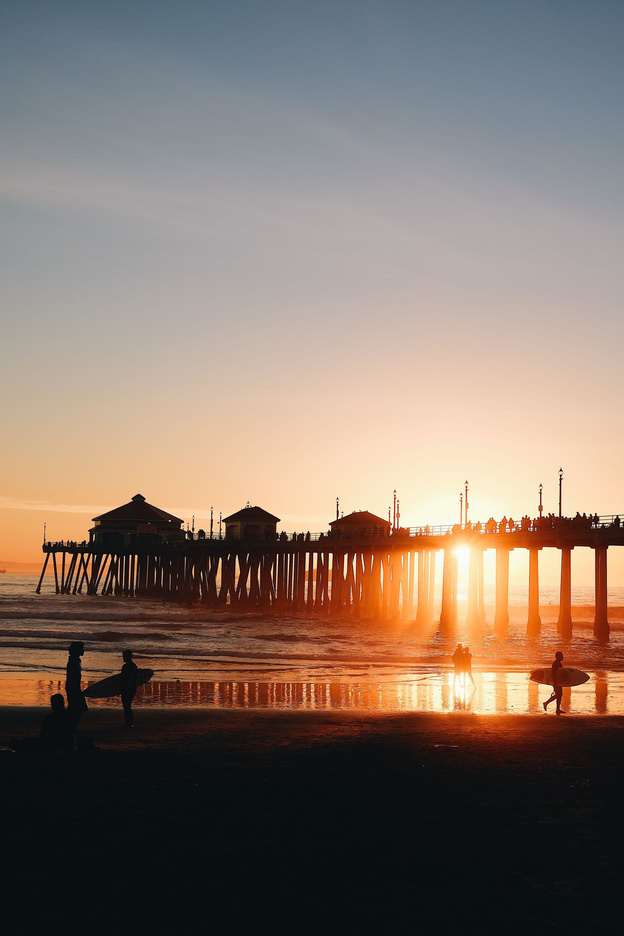 Surfers walking the sand near the Huntington Beach pier at sunset.